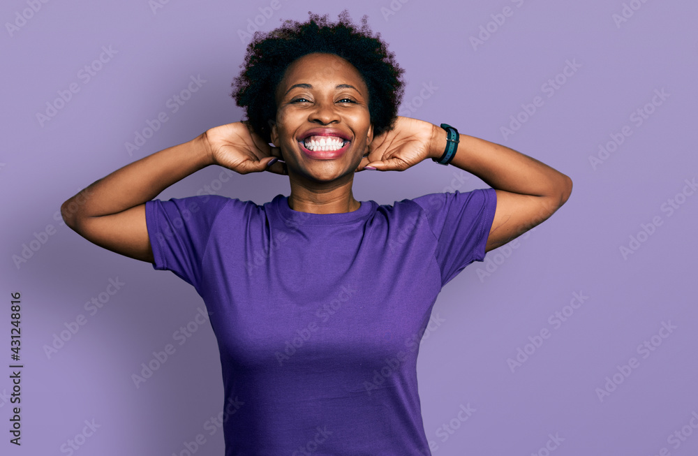 African american woman with afro hair wearing casual purple t shirt relaxing and stretching, arms and hands behind head and neck smiling happy