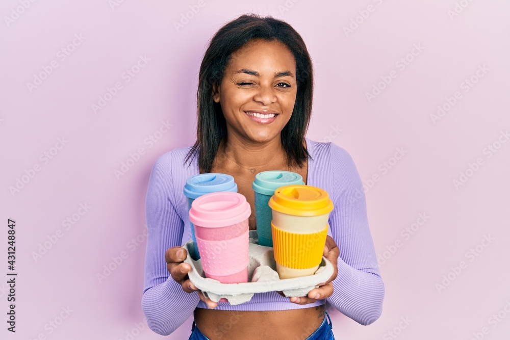 Young african american girl holding tray with take away coffee winking looking at the camera with sexy expression, cheerful and happy face. Young african american girl holding tray with take away coffee winking looking at the camera with sexy expression, cheerful and happy face.