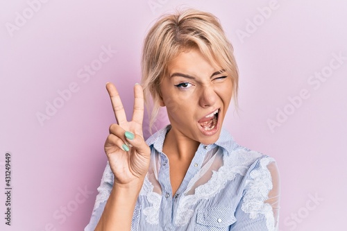 Young blonde girl wearing casual clothes smiling with happy face winking at the camera doing victory sign. number two.