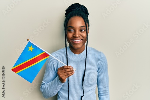 African american woman with braided hair holding democratic republic of the congo flag looking positive and happy standing and smiling with a confident smile showing teeth