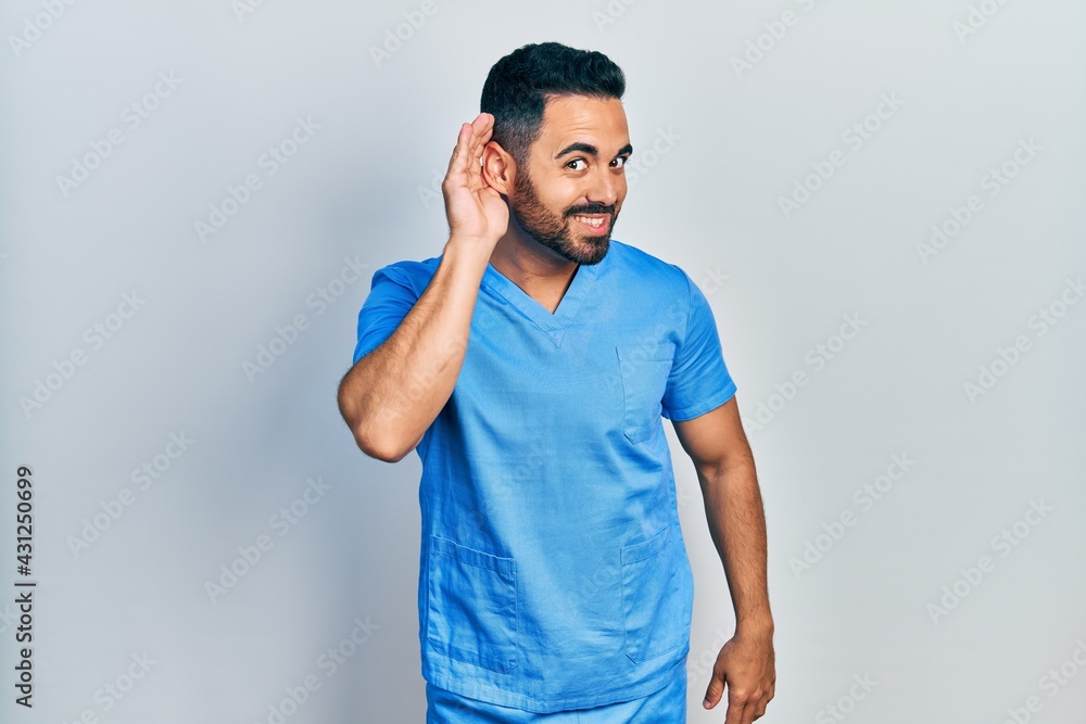 Handsome hispanic man with beard wearing blue male nurse uniform smiling with hand over ear listening an hearing to rumor or gossip. deafness concept. Handsome hispanic man with beard wearing blue male nurse uniform smiling with hand over ear listening an hearing to rumor or gossip. deafness concept.