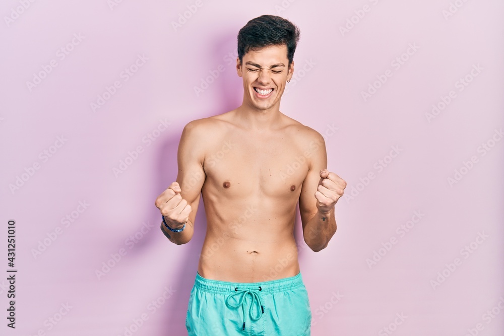 Fototapeta premium Young hispanic man wearing swimwear shirtless very happy and excited doing winner gesture with arms raised, smiling and screaming for success. celebration concept.