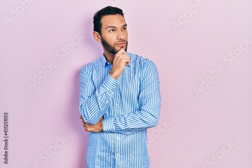 Hispanic man with beard wearing casual blue shirt with hand on chin thinking about question, pensive expression. smiling with thoughtful face. doubt concept.