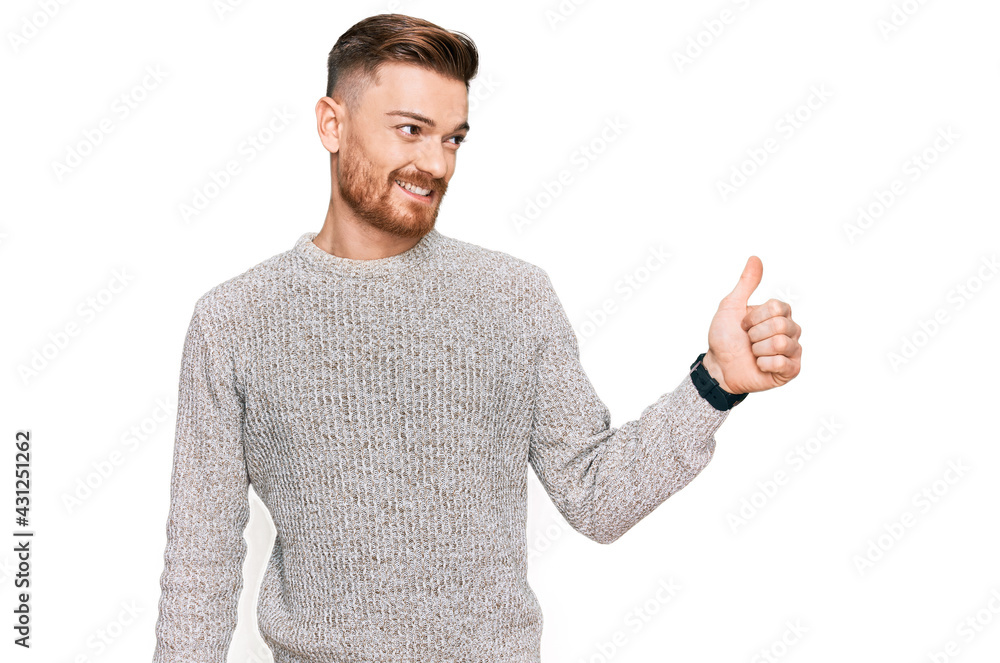 Young redhead man wearing casual winter sweater looking proud, smiling doing thumbs up gesture to the side Young redhead man wearing casual winter sweater looking proud, smiling doing thumbs up gesture to the side