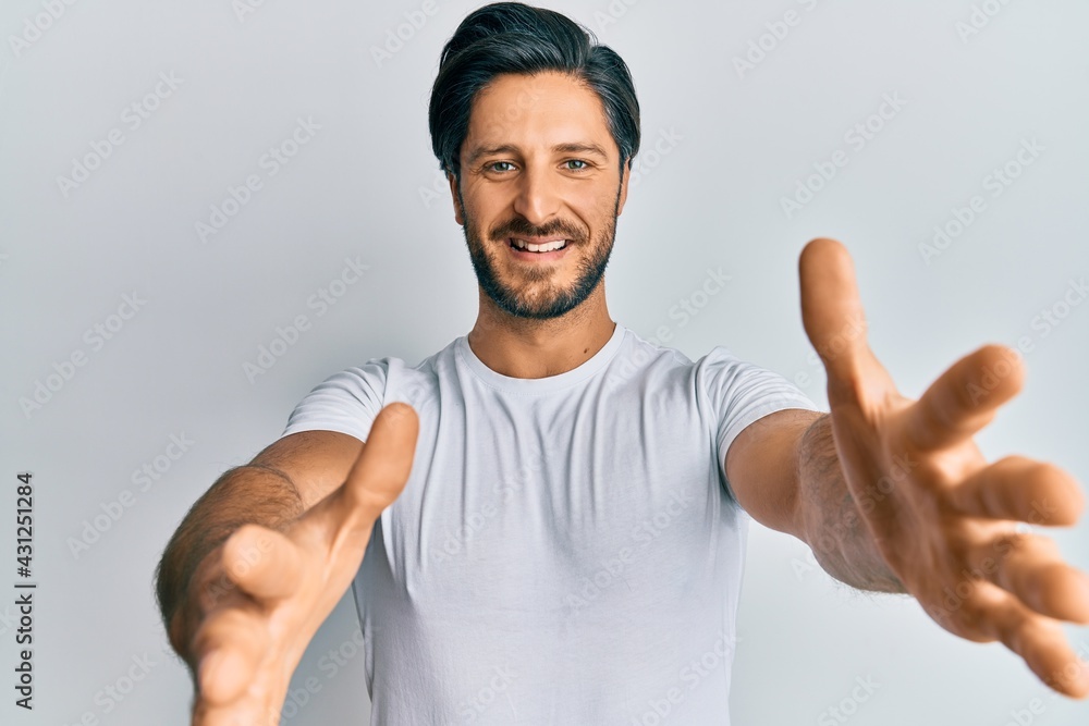 Young hispanic man wearing casual white t shirt looking at the camera smiling with open arms for hug. cheerful expression embracing happiness. Young hispanic man wearing casual white t shirt looking at the camera smiling with open arms for hug. cheerful expression embracing happiness.