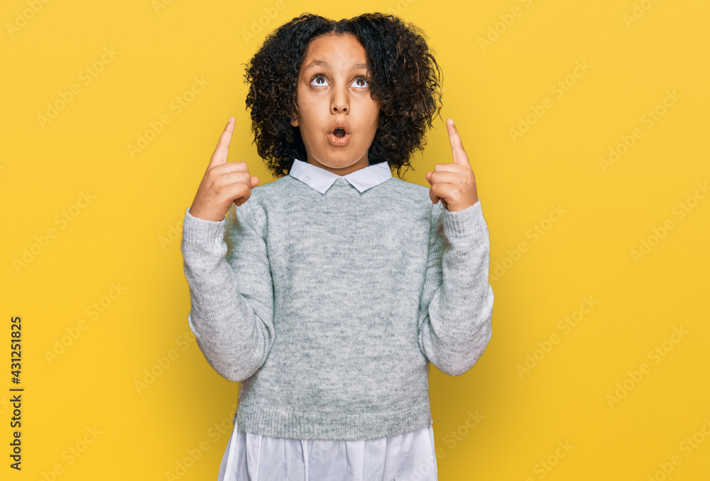 Young little girl with afro hair wearing casual clothes amazed and surprised looking up and pointing with fingers and raised arms. Young little girl with afro hair wearing casual clothes amazed and surprised looking up and pointing with fingers and raised arms.