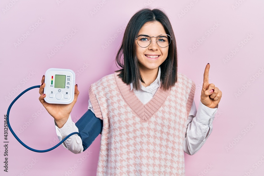 Young brunette woman using blood pressure monitor smiling with an idea or question pointing finger with happy face, number one Young brunette woman using blood pressure monitor smiling with an idea or question pointing finger with happy face, number one