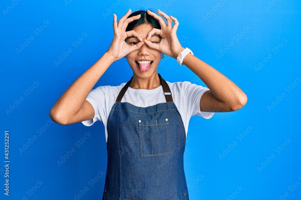Young chinese woman wearing waiter apron doing ok gesture like binoculars sticking tongue out, eyes looking through fingers. crazy expression. Young chinese woman wearing waiter apron doing ok gesture like binoculars sticking tongue out, eyes looking through fingers. crazy expression.