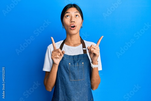 Young chinese woman wearing waiter apron amazed and surprised looking up and pointing with fingers and raised arms.