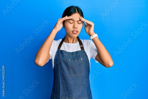 Young chinese woman wearing waiter apron with hand on head for pain in head because stress. suffering migraine.