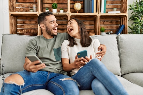 Young latin couple using smartphone sitting on the sofa at home.