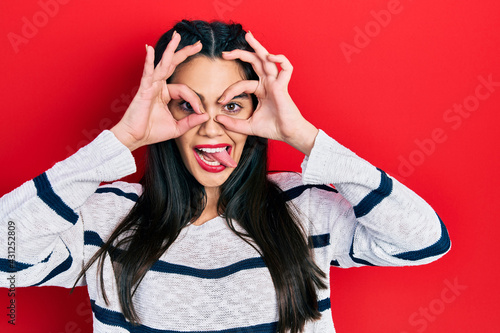 Young hispanic girl wearing casual clothes doing ok gesture like binoculars sticking tongue out, eyes looking through fingers. crazy expression.