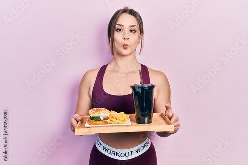 Young hispanic girl wearing sportswear eating a tasty classic burger with fries and soda making fish face with mouth and squinting eyes, crazy and comical.
