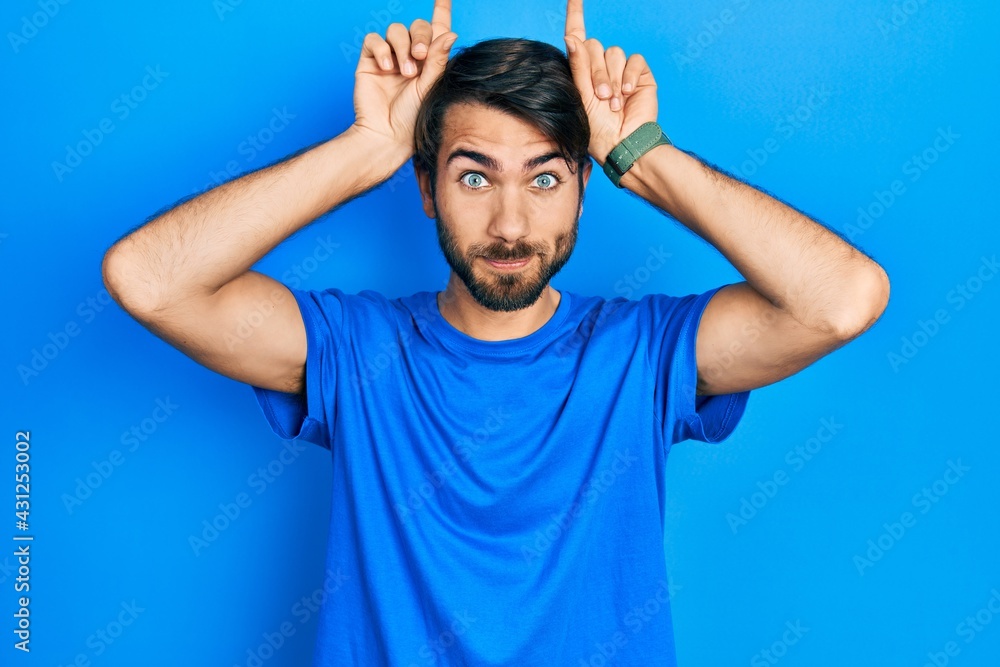 Young hispanic man wearing casual clothes doing funny gesture with finger over head as bull horns Young hispanic man wearing casual clothes doing funny gesture with finger over head as bull horns