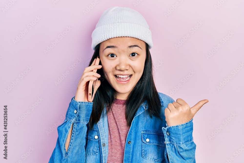 Young chinese girl having conversation talking on the smartphone pointing thumb up to the side smiling happy with open mouth Young chinese girl having conversation talking on the smartphone pointing thumb up to the side smiling happy with open mouth
