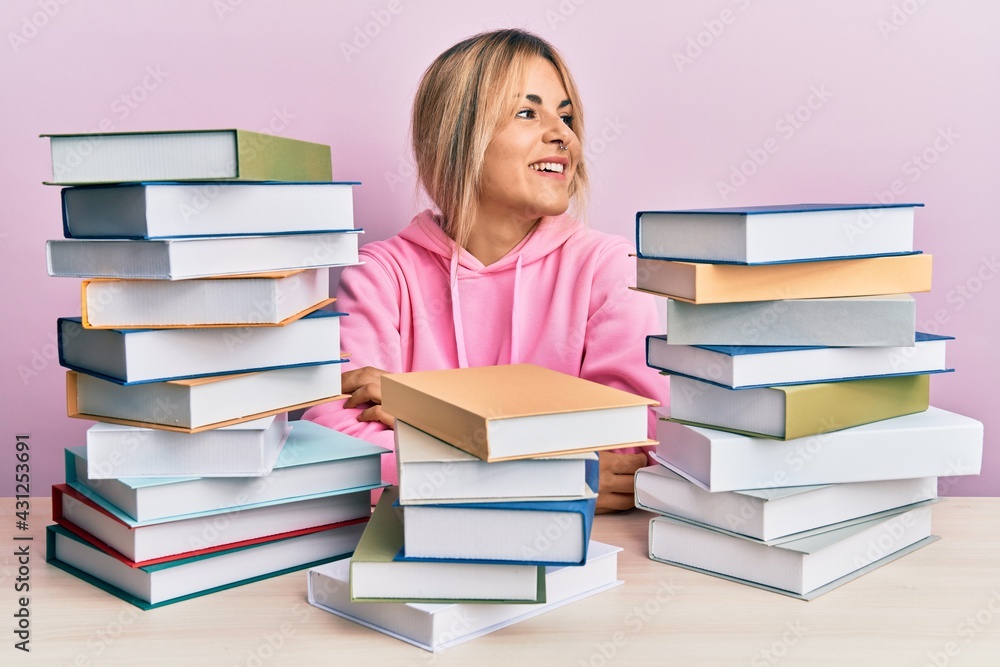 Young caucasian woman sitting on the table with books looking away to side with smile on face, natural expression. laughing confident. Young caucasian woman sitting on the table with books looking away to side with smile on face, natural expression. laughing confident.