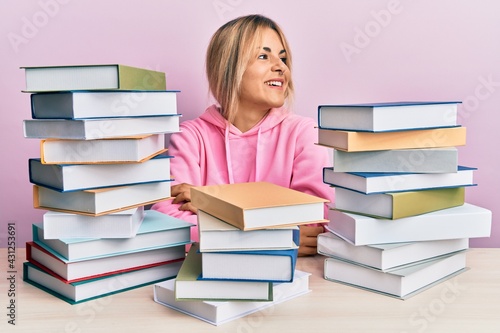 Young caucasian woman sitting on the table with books looking away to side with smile on face, natural expression. laughing confident.