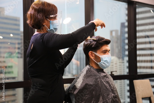 Barber and customer wearing facial mask in hair salon while having a haircut for avoiding contagion during new normal and social distancing lifestyle after coronavirus or covid-19 pandemic