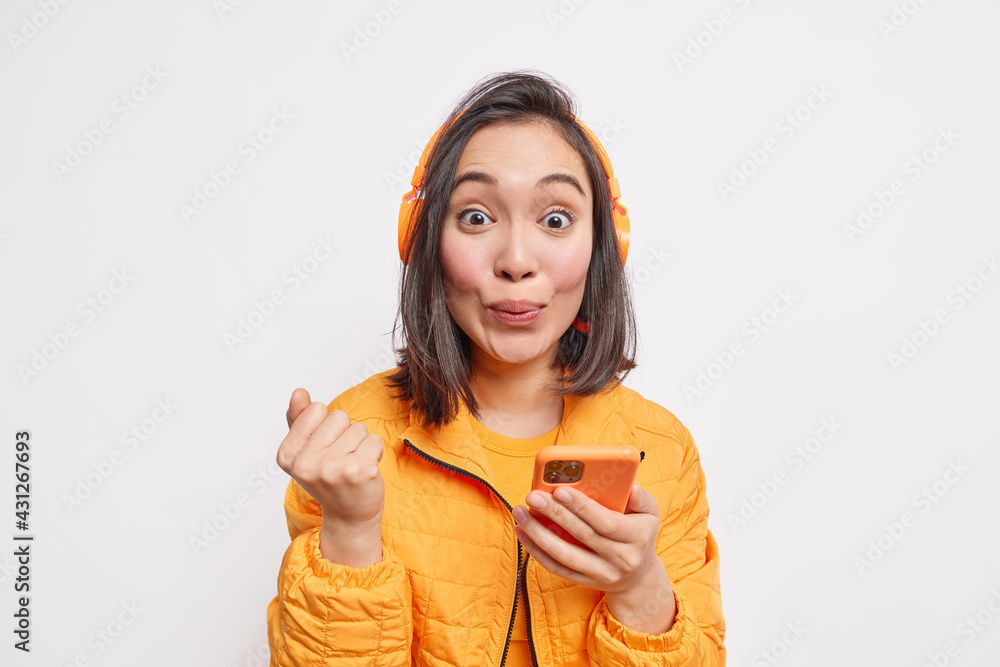 Photo of good looking Asian woman with dark hair clenches fist uses smartphone for online communication gets new music subscription dressed in orange jacket isolated over white studio background Photo of good looking Asian woman with dark hair clenches fist uses smartphone for online communication gets new music subscription dressed in orange jacket isolated over white studio background