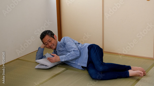 和室で寝転がってスマホを操作するシニア男性 © Ameashi