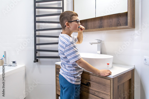 A little boy with glasses in the bathroom brushes his teeth.