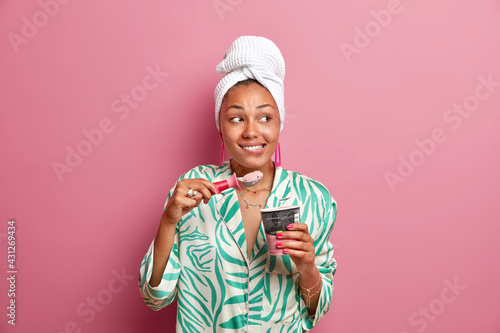 Indoor shot of cheerful dark skinned young woman bites lips enjoys eating appetizing cold ice cream from spoon spends free time at home dressed in casual clothes isolated over pink background