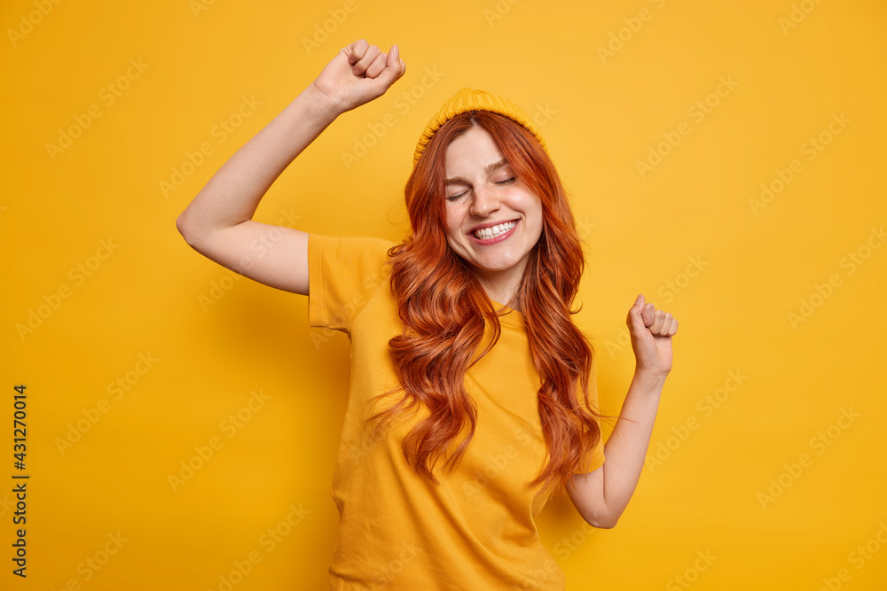 Studio shot of carefree redhead woman raises hand dances and smiles cheerfully has fun wears hat and casual t shirt isolated over vivid yellow background partying in good mood. Monochrome shot. Studio shot of carefree redhead woman raises hand dances and smiles cheerfully has fun wears hat and casual t shirt isolated over vivid yellow background partying in good mood. Monochrome shot.