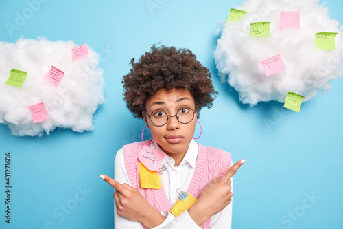 Puzzled hesitant skilled female business worker poses around colorful reminder stickers hesitates between two options wears round spectacles isolated over blue background white clouds overhead photo