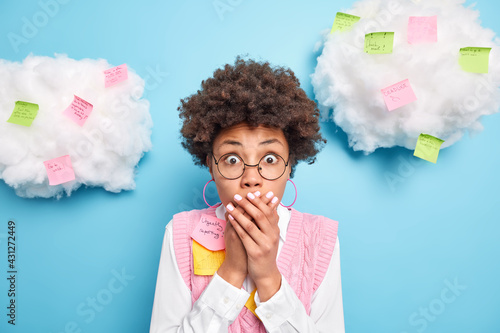 Indoor shot of shocked ethnic female office worker keeps hands on mouth writes tasks and creative ideas on sticky post it notes realises deadline wears round spectacles formal clothes stands indoor photo