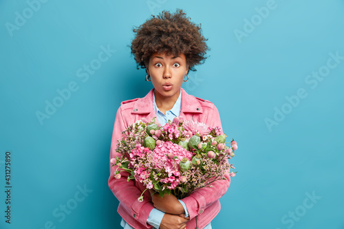 Photo of surprised young African American woman with curly hair embraces big nice bunch of flowers shocked to get congratulations on birthday isolated over blue background. Festive event concept