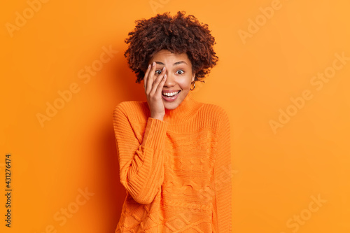 Portrait of happy woman with curly bushy hair enjoys leisure free time makes face palm smiles broadly being in good mood wears casual bright jumper isolated over orange background. Emotions concept © Wayhome Studio