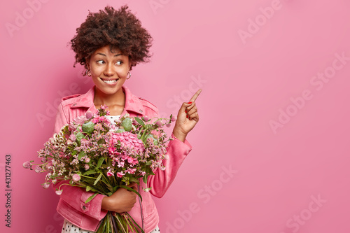 Positive dark skinned young curly woman poses with nice bouquet of flowers points at blank space shows advertising content wears jacket isolated over pink background. Happy Mothers Day concept
