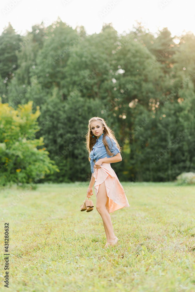 A beautiful young woman holds summer shoes in her hands and walks barefoot on the grass on a summer day A beautiful young woman holds summer shoes in her hands and walks barefoot on the grass on a summer day
