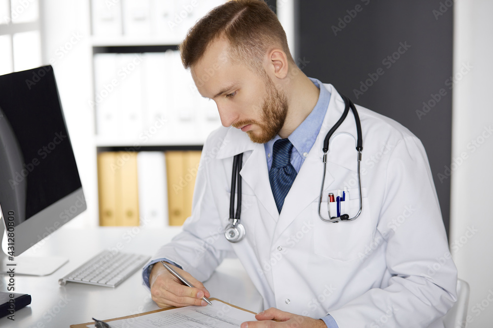 Friendly red-bearded doctor sitting and writing at clipboard in clinic. Perfect medical service in hospital. Medicine, healthcare Friendly red-bearded doctor sitting and writing at clipboard in clinic. Perfect medical service in hospital. Medicine, healthcare