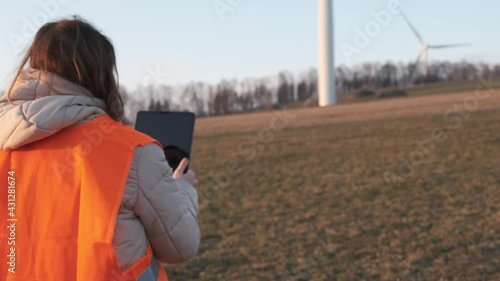 Female engineer in orange vesta goes to wind turbines with a tablet to checks their operation. 