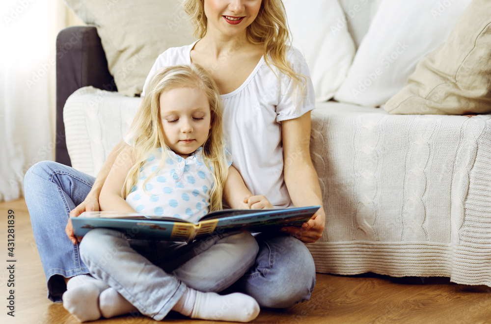 Happy family. Blonde young mother reading a book to her cute daughter while sitting at wooden floor. Motherhood concept Happy family. Blonde young mother reading a book to her cute daughter while sitting at wooden floor. Motherhood concept