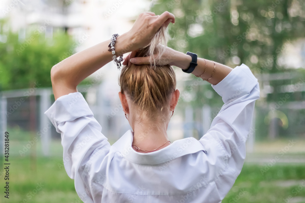 The blonde girl stands with her back to the camera and pulls her hair into a ponytail.