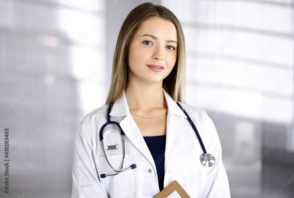 Young optimistic woman-doctor is holding a clipboard in her hands, while standing in a clinic. Portrait of friendly female physician with a stethoscope. Perfect medical service in a hospital. Medicine Young optimistic woman-doctor is holding a clipboard in her hands, while standing in a clinic. Portrait of friendly female physician with a stethoscope. Perfect medical service in a hospital. Medicine