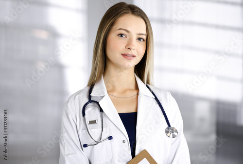 Young optimistic woman-doctor is holding a clipboard in her hands, while standing in a clinic. Portrait of friendly female physician with a stethoscope. Perfect medical service in a hospital. Medicine