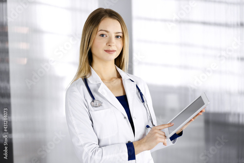 Young smiling woman-doctor is holding a tablet computer in her hands, while standing in the cabinet in a clinic. Portrait of friendly female physician with a stethoscope. Perfect medical service in a