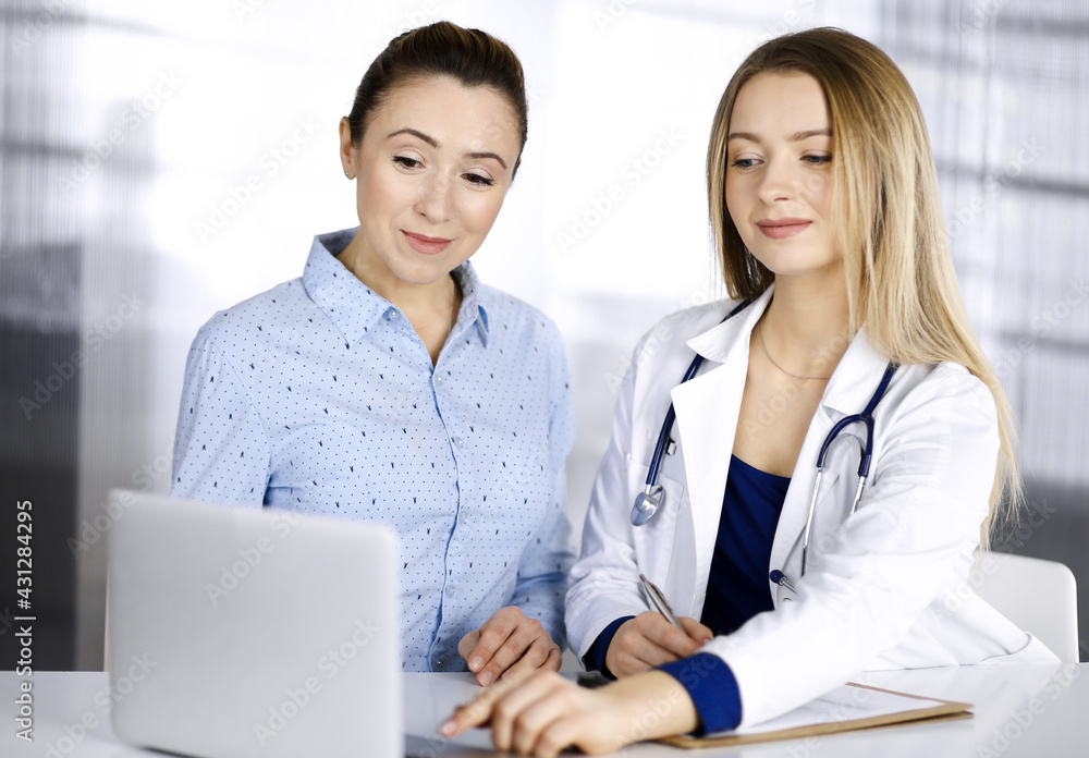 Young beautiful woman-doctor and her patient are discussing patient's current health examination, while sitting together at the desk in the cabinet in a clinic. Female physician is writing some notes Young beautiful woman-doctor and her patient are discussing patient's current health examination, while sitting together at the desk in the cabinet in a clinic. Female physician is writing some notes