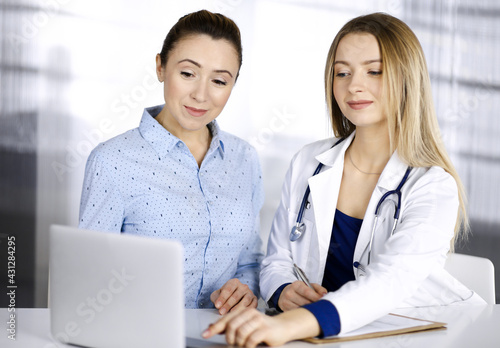 Young beautiful woman-doctor and her patient are discussing patient's current health examination, while sitting together at the desk in the cabinet in a clinic. Female physician is writing some notes