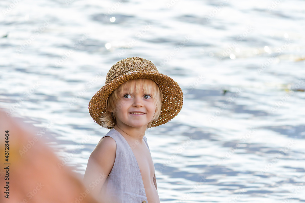 Portrait of little happy caucasian blond boy wearing straw hat against river water in the background. Summer kids fun theme.