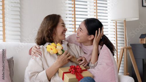 Attractive beautiful asian middle age mum sit with grown up daughter give gift box and flower in family moment celebrate mother day. Overjoy bonding cheerful kid embrace relationship with retired mom.
