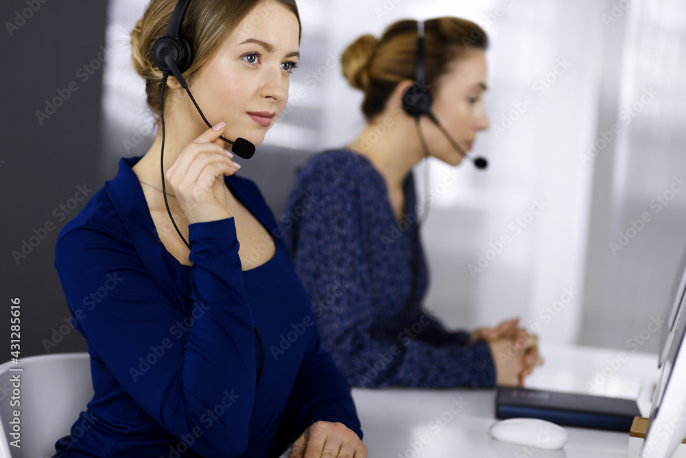 Two busineswomen have conversations with the clients by headsets, while sitting at the desk in a modern office. Diverse people group in a call center. Telemarketing and customer service Two busineswomen have conversations with the clients by headsets, while sitting at the desk in a modern office. Diverse people group in a call center. Telemarketing and customer service