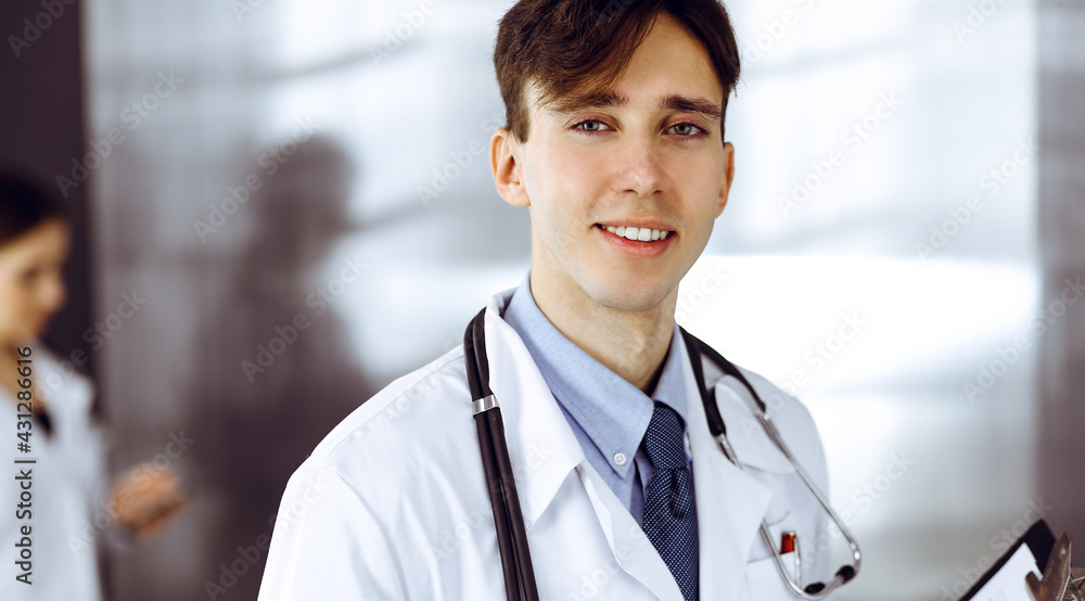 Friendly young male doctor standing with clipboard in modern clinic. Female colleague is busy with filling up of medication records form at the background of physician. Medicine concept Friendly young male doctor standing with clipboard in modern clinic. Female colleague is busy with filling up of medication records form at the background of physician. Medicine concept