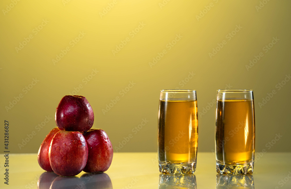 Two glasses of apple juice with red apples in the sunset background. yellow background. Reflected surface. Summer day. Good mood. Two glasses of apple juice with red apples in the sunset background. yellow background. Reflected surface. Summer day. Good mood.