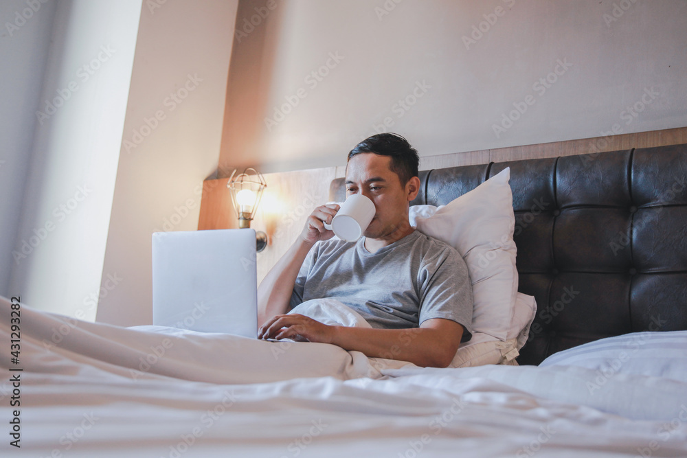 Asian man work from home by using laptop on the bed while drink a coffee Asian man work from home by using laptop on the bed while drink a coffee