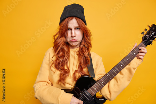 Distressed unhappy redhead young woman plays bass electric guitar has sad expression wears black hat and casual yellow sweatshirt poses indoor. Displeased female rocker with musical instrument