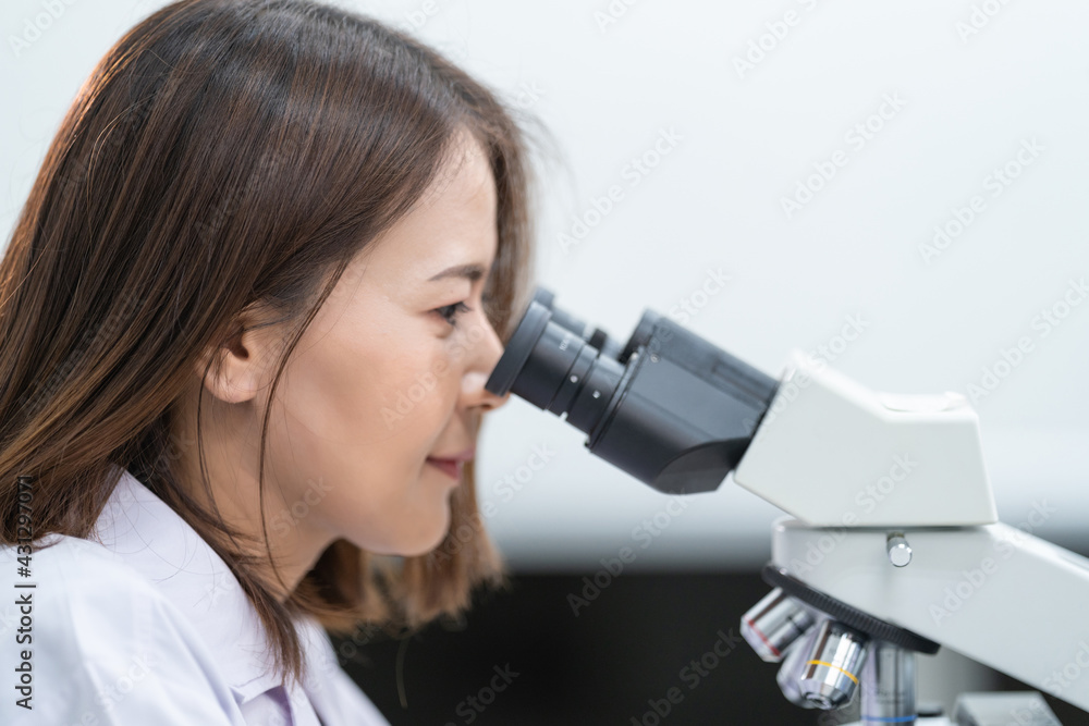 A young scientist woman in a laboratory coat looking through a microscope in a laboratory to do research and experiment. Scientist working in a laboratory. Education stock photo A young scientist woman in a laboratory coat looking through a microscope in a laboratory to do research and experiment. Scientist working in a laboratory. Education stock photo